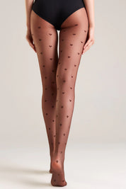 A model from the waist down wearing sheer brown hosiery with a small dark heart pattern, paired with black underwear, against a light gray studio background.