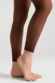 A close-up of a person's legs wearing sheer, dark brown tights against a clean, light gray studio background.
