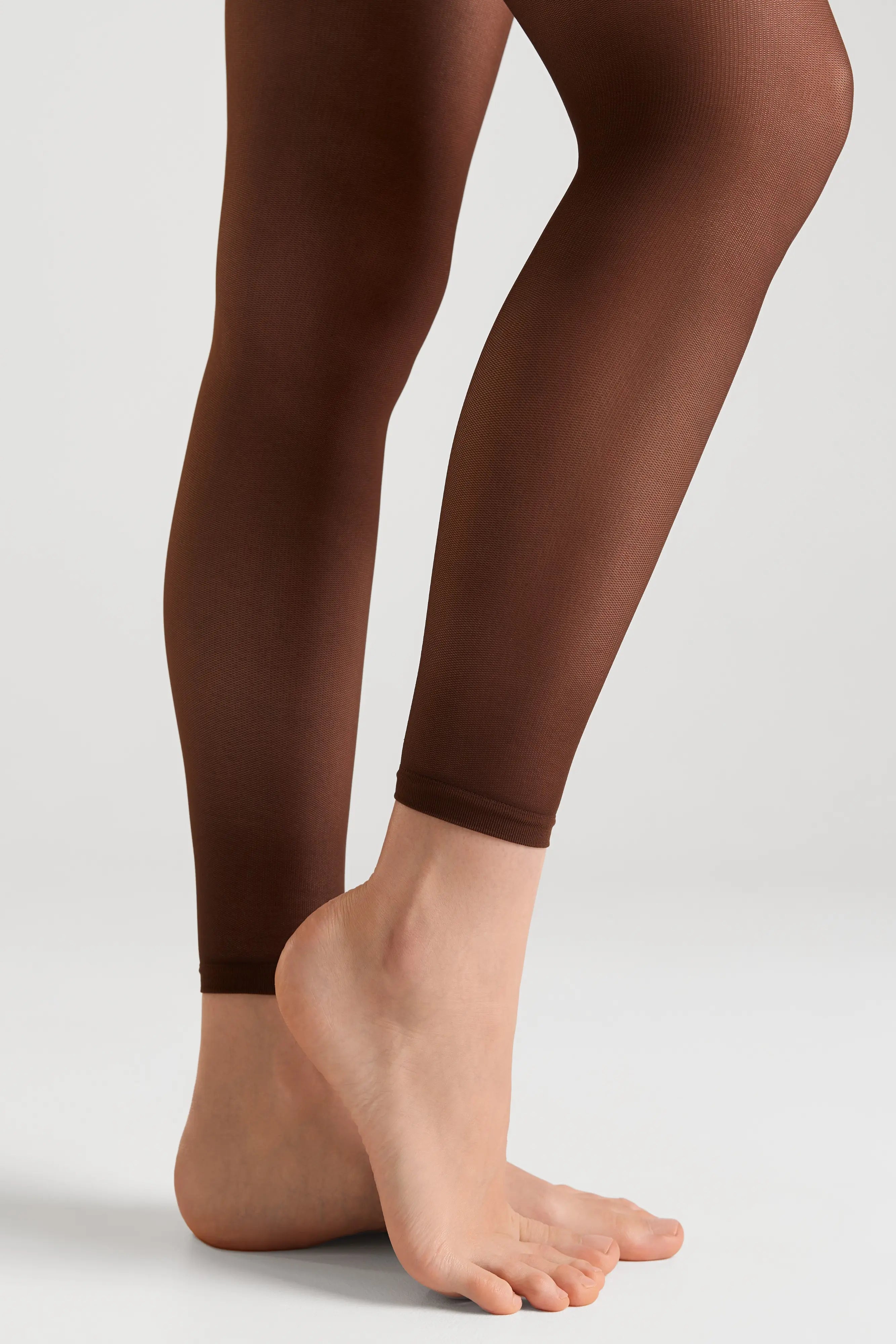 A close-up of a person's legs wearing sheer, dark brown tights against a clean, light gray studio background.