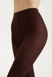 A close-up side view of a model wearing high-waisted, solid dark brown tights against a clean, light off-white background.