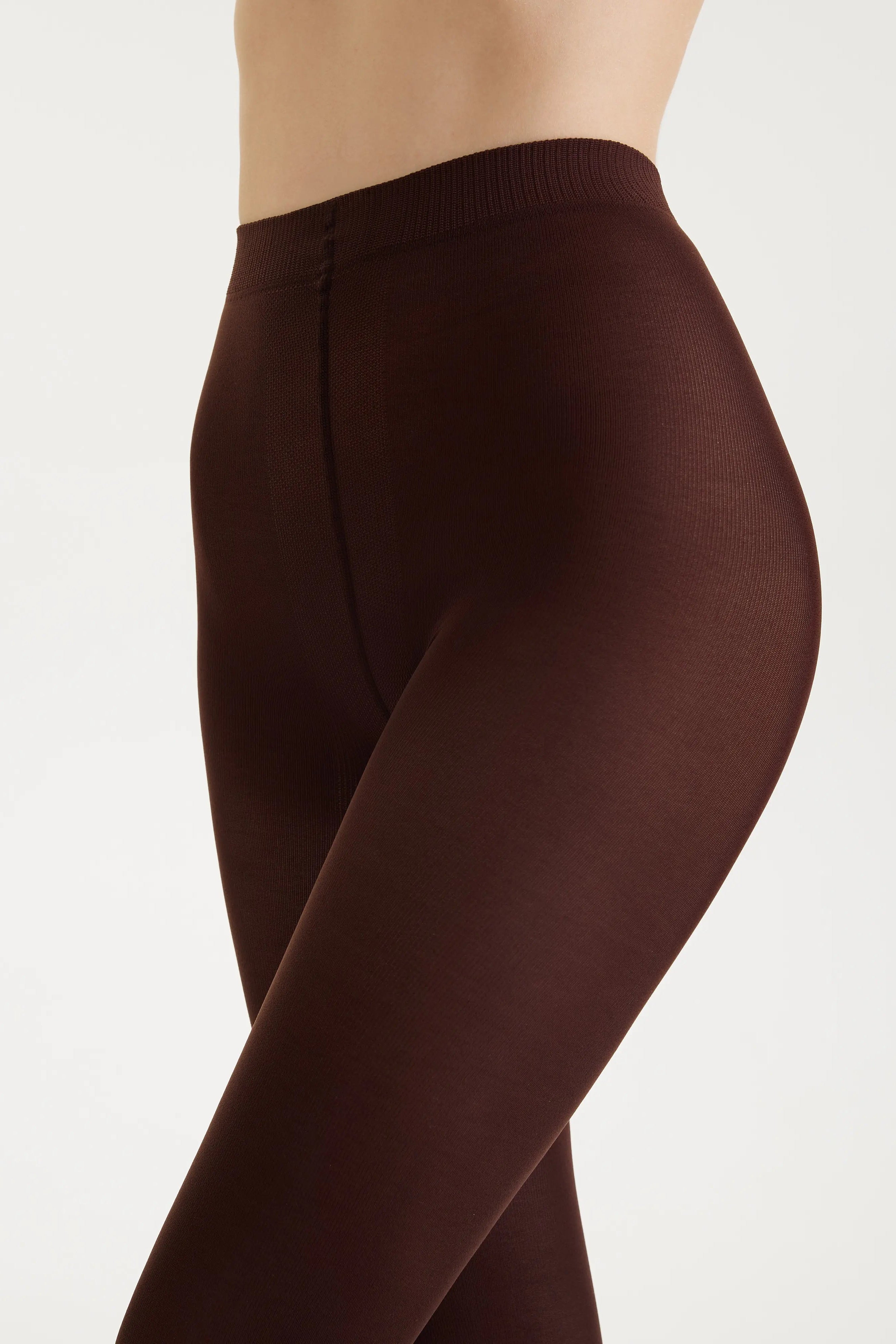 A close-up side view of a model wearing high-waisted, solid dark brown tights against a clean, light off-white background.