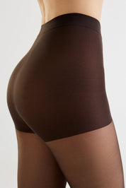 Close-up side view of a model wearing high-waisted brown shapewear briefs and sheer brown tights against a light gray studio background.