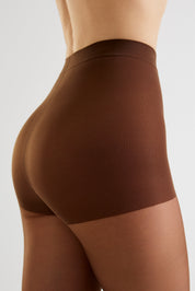 Close-up side view of a model wearing high-waisted brown compression shorts or shapewear against a light gray studio background.