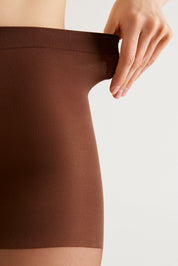 A close-up of a person wearing a brown ribbed compression or shapewear brief, with their hand stretching the elastic waistband against a plain light background.