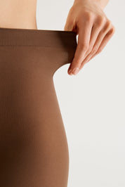 Close-up of a person wearing a ribbed brown hosiery stocking, pulling the waistband slightly outward with a hand, against a clean off-white background.