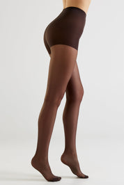 Side view of a model wearing sheer dark brown tights with a matching opaque waistband, shown against a plain light gray studio background.