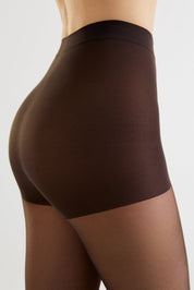 Close-up side view of a model wearing high-waisted brown shapewear briefs and sheer brown tights against a light gray studio background.