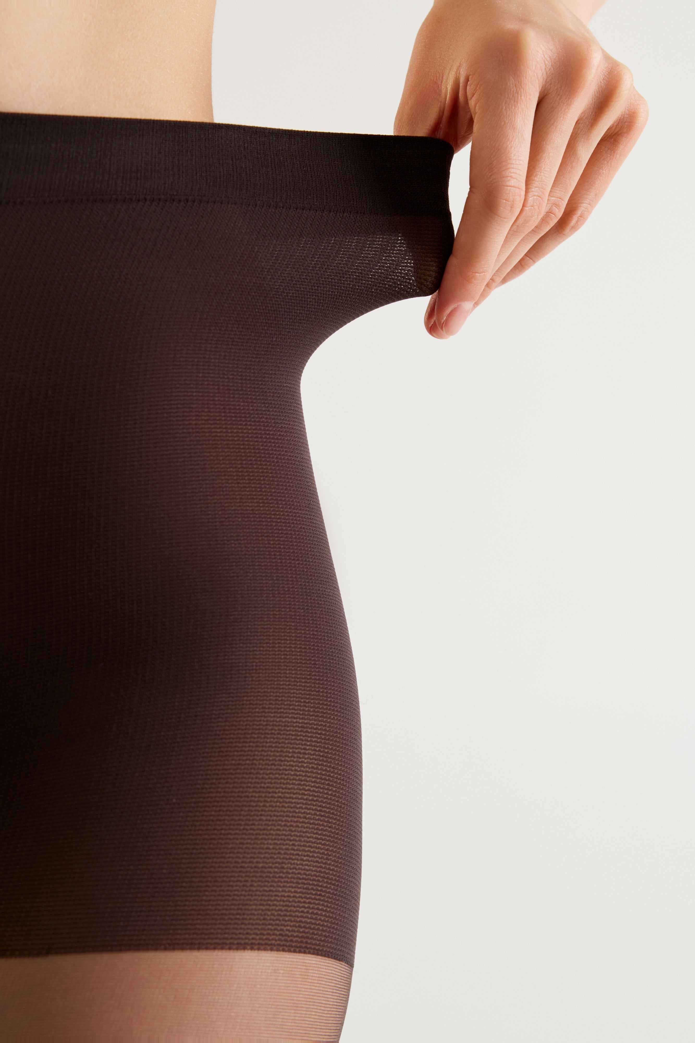 Close-up of a person wearing dark brown ribbed compression shorts or a thigh-high stocking, with a hand stretching the waistband to show elasticity, against a light gray background.
