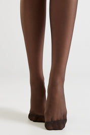 A close-up of a person's legs wearing sheer dark brown tights against a plain light gray studio background.