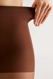 Close-up of a person wearing a brown ribbed shapewear brief or waist trainer, with one hand lifting the waistband against a plain light background.