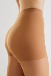 Close-up side view of a model wearing high-waisted ribbed shapewear briefs in a warm nude tan color against a light gray studio background.