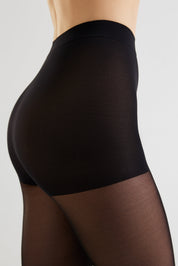Close-up side view of a model wearing black high-waisted compression hosiery with sheer black tights visible below, against a light gray studio background.