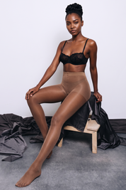A model wearing sheer nude tights, a black lace bralette, and beige shapewear briefs sits on a small wooden stool draped with black fabric against a light gray studio background.