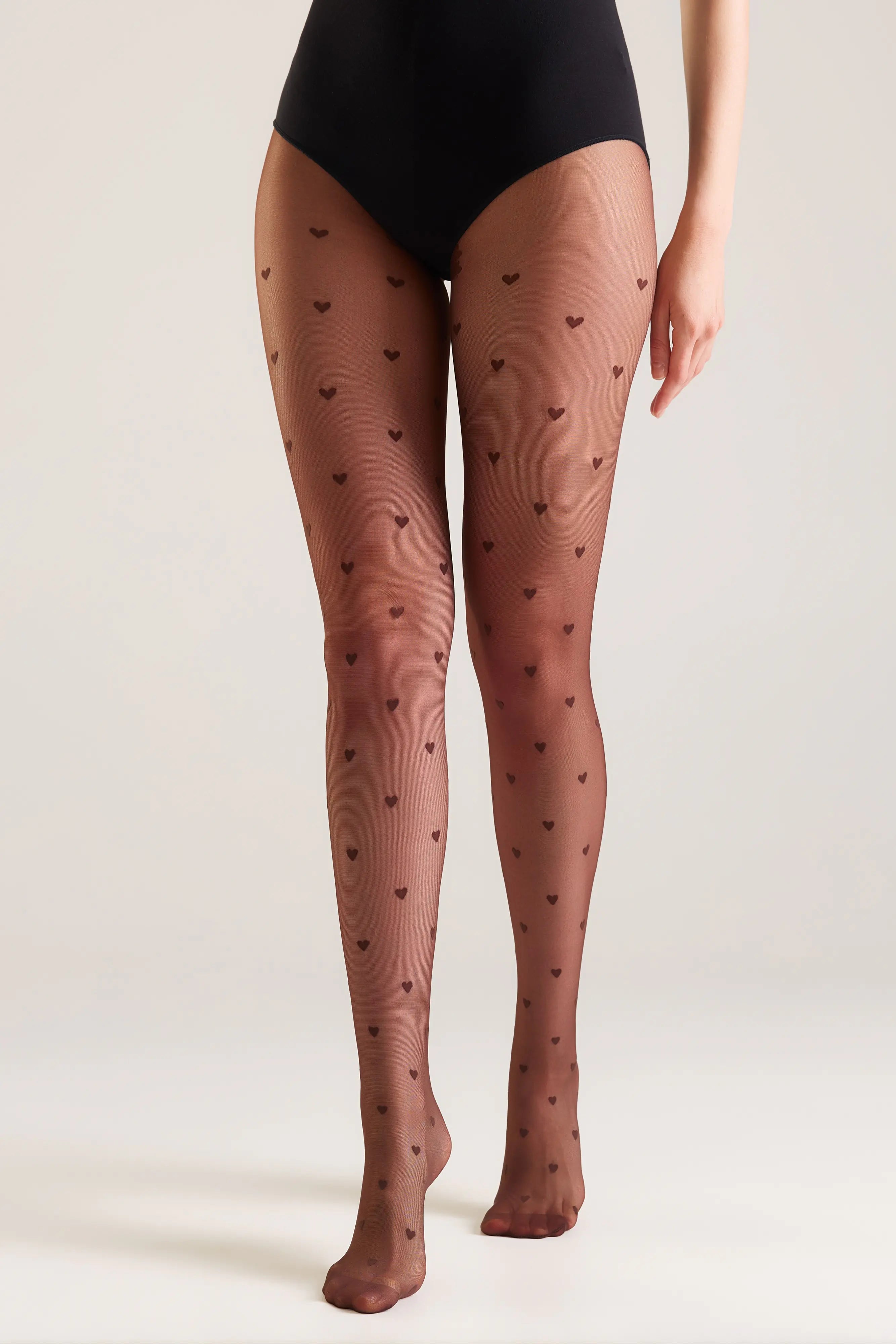 A model from the waist down wearing sheer black tights with a small repeating heart pattern, paired with a black bodysuit, against a light gray studio background.