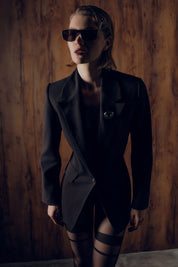 Woman wearing a black coat and sunglasses against a wooden wall.