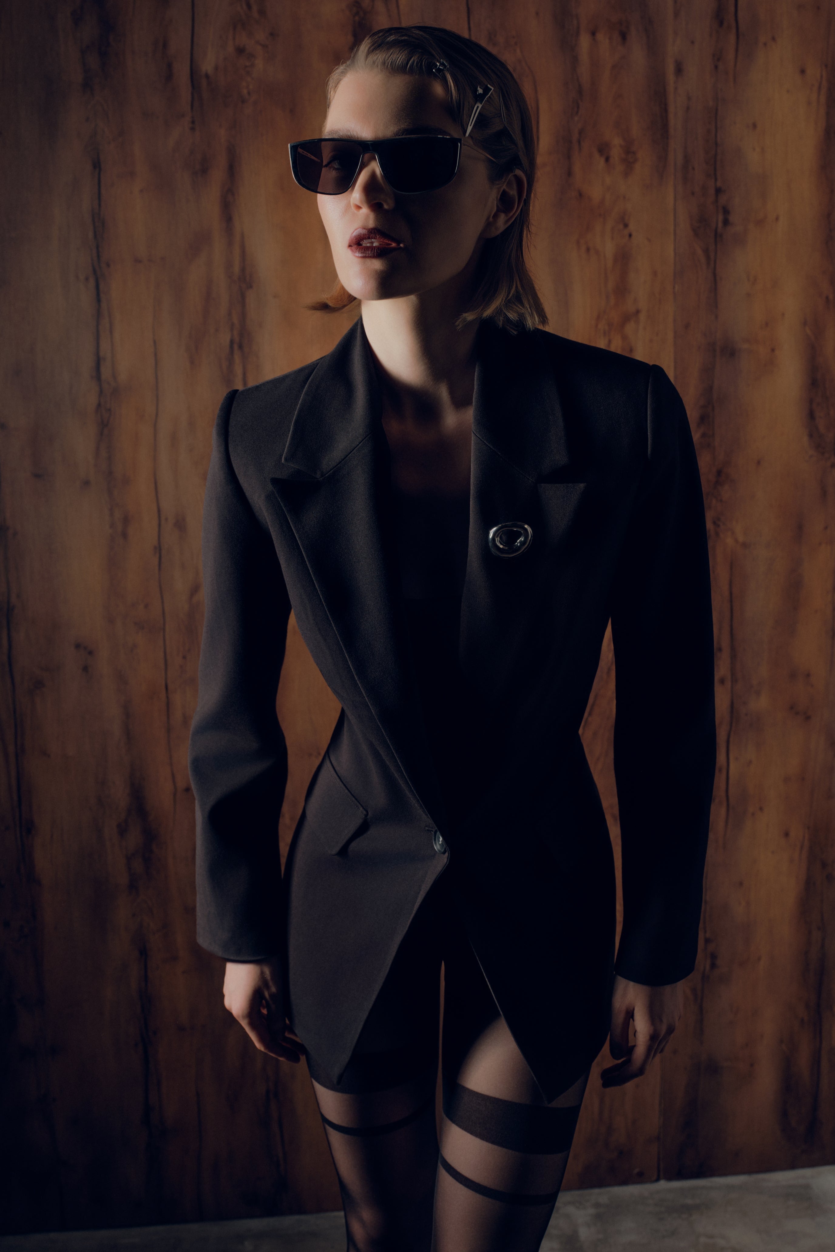 Woman wearing a black coat and sunglasses against a wooden wall.