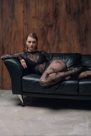Woman in black lace outfit sitting on a black leather couch against a wooden wall.