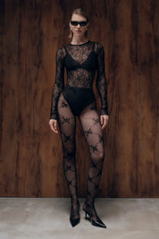 Model wearing a black lace bodysuit and stockings against a wooden background