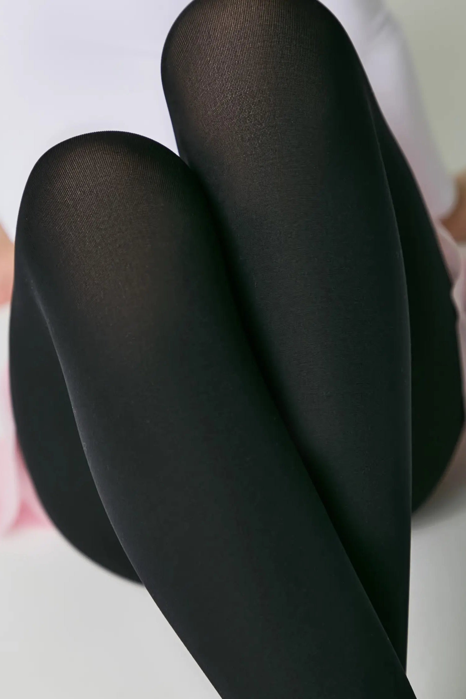 Black tights worn by a person sitting on a pink surface with a white background #color_nero-black