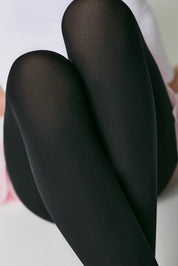 Black tights worn by a person sitting on a pink surface with a white background #color_nero-black
