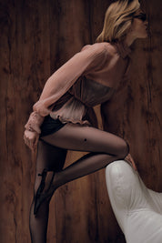 Woman in a sheer blouse and black stockings sitting on a white chair against a wooden wall.