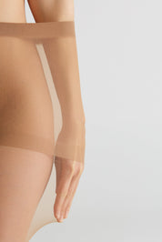 Close-up of a model's leg and hand wearing sheer nude thigh-high stockings against a light gray background.