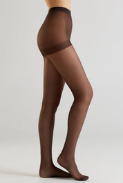 A model's lower body wearing sheer dark brown hosiery with a wide opaque brown waistband, shown against a light gray studio background.