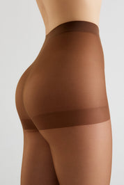 Close-up side view of a model wearing sheer brown compression hosiery with a wide supportive band at the upper thigh, shown against a light gray studio background.