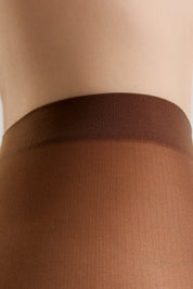 Close-up of a person's lower torso wearing sheer brown hosiery with a wide ribbed waistband, against a light neutral background.