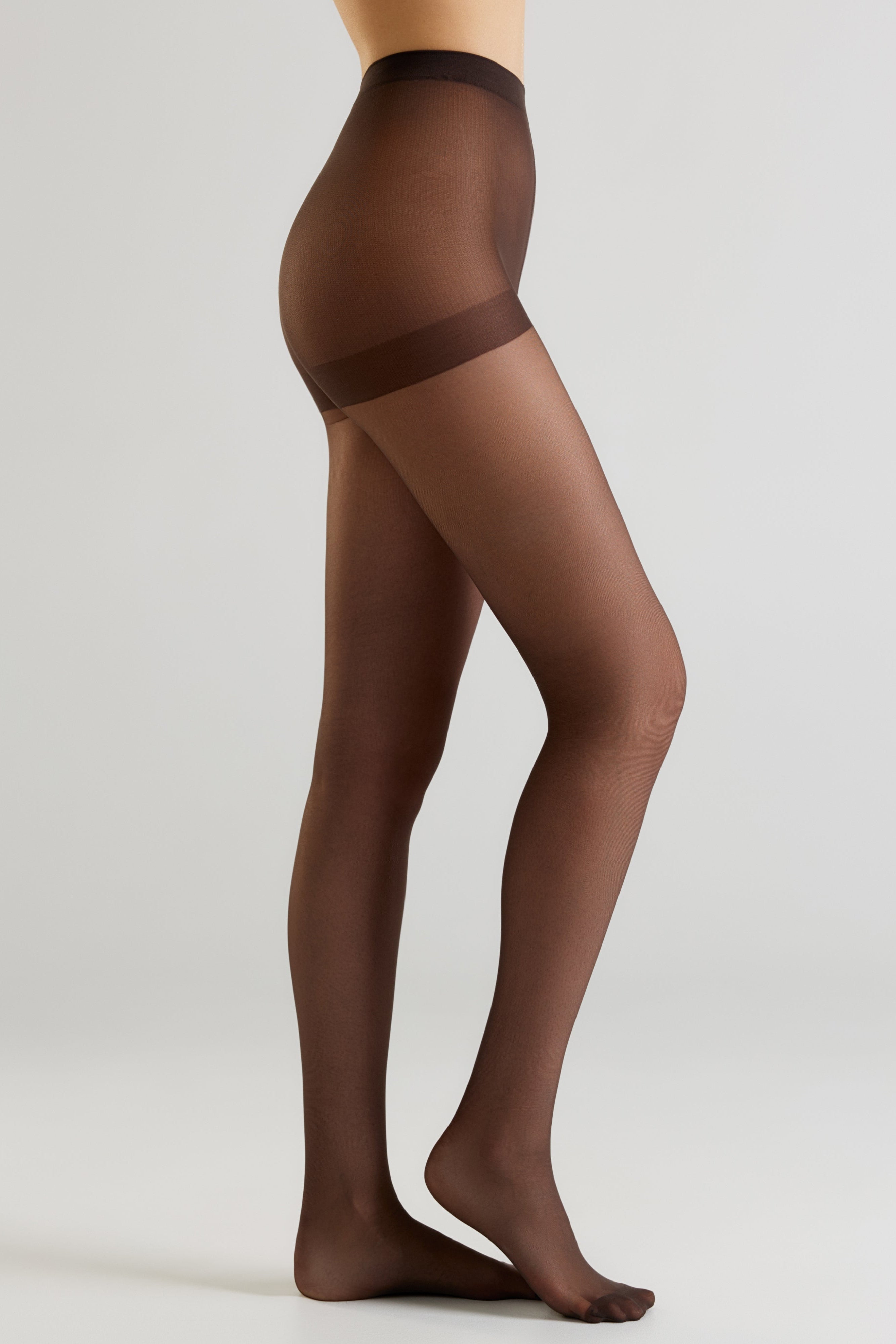 A model's lower body wearing sheer dark brown hosiery with a wide opaque brown waistband, shown against a light gray studio background.