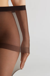 Close-up of a model wearing sheer thigh-high stockings in a dark brown shade, showing the leg and hand against a light gray studio background.