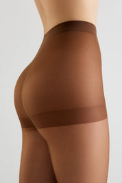 Close-up side view of a model wearing sheer brown compression hosiery with a wide supportive band at the upper thigh, shown against a light gray studio background.