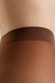 Close-up of a person's lower torso wearing sheer brown hosiery with a wide ribbed waistband, against a light neutral background.