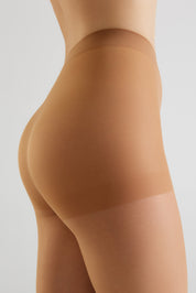 Close-up side view of a model wearing sheer beige hosiery or shapewear on the hip and upper thigh, against a light gray studio background.