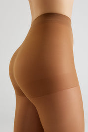 Close-up side view of a model wearing sheer, high-waisted hosiery in a warm tan shade against a light gray studio background.