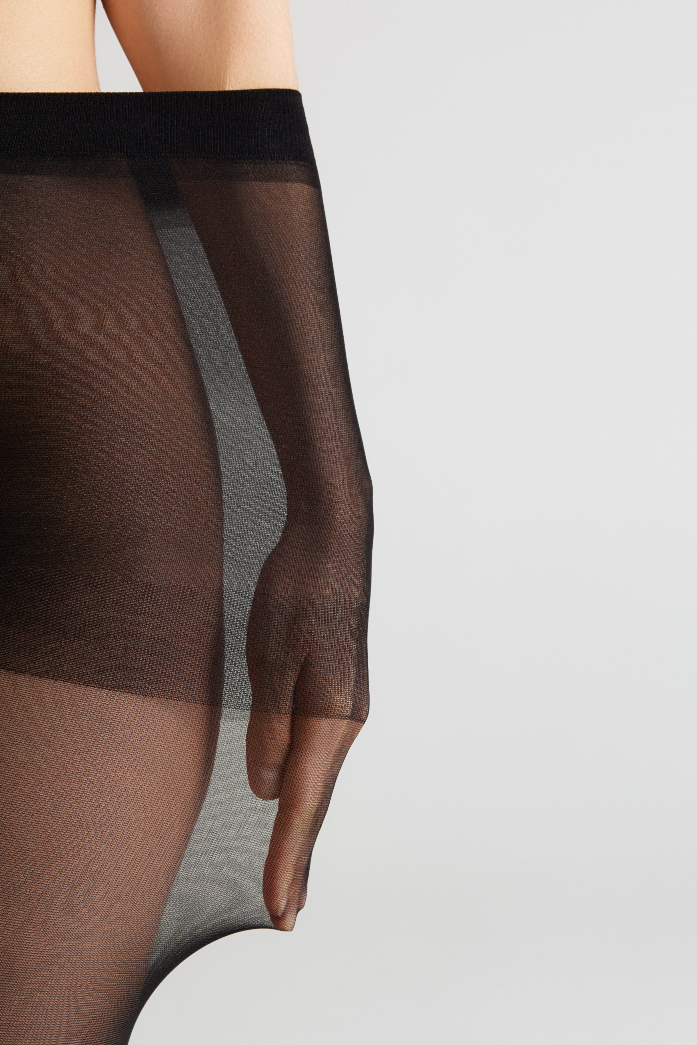 Close-up of a model wearing sheer black thigh-high stockings, showing the semi-transparent fabric and waistband against a light gray studio background.