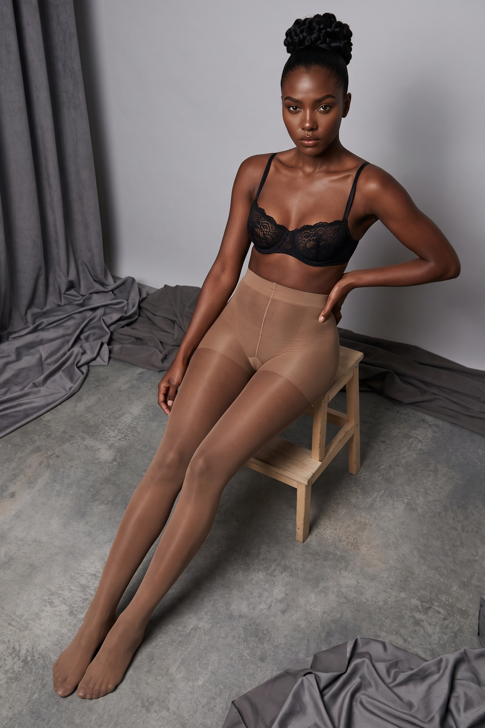A model sits on a small wooden stool in a studio setting, wearing sheer nude tights, a black lace bralette, and high-waisted beige shapewear briefs. The background features a gray wall and floor with draped gray fabric.