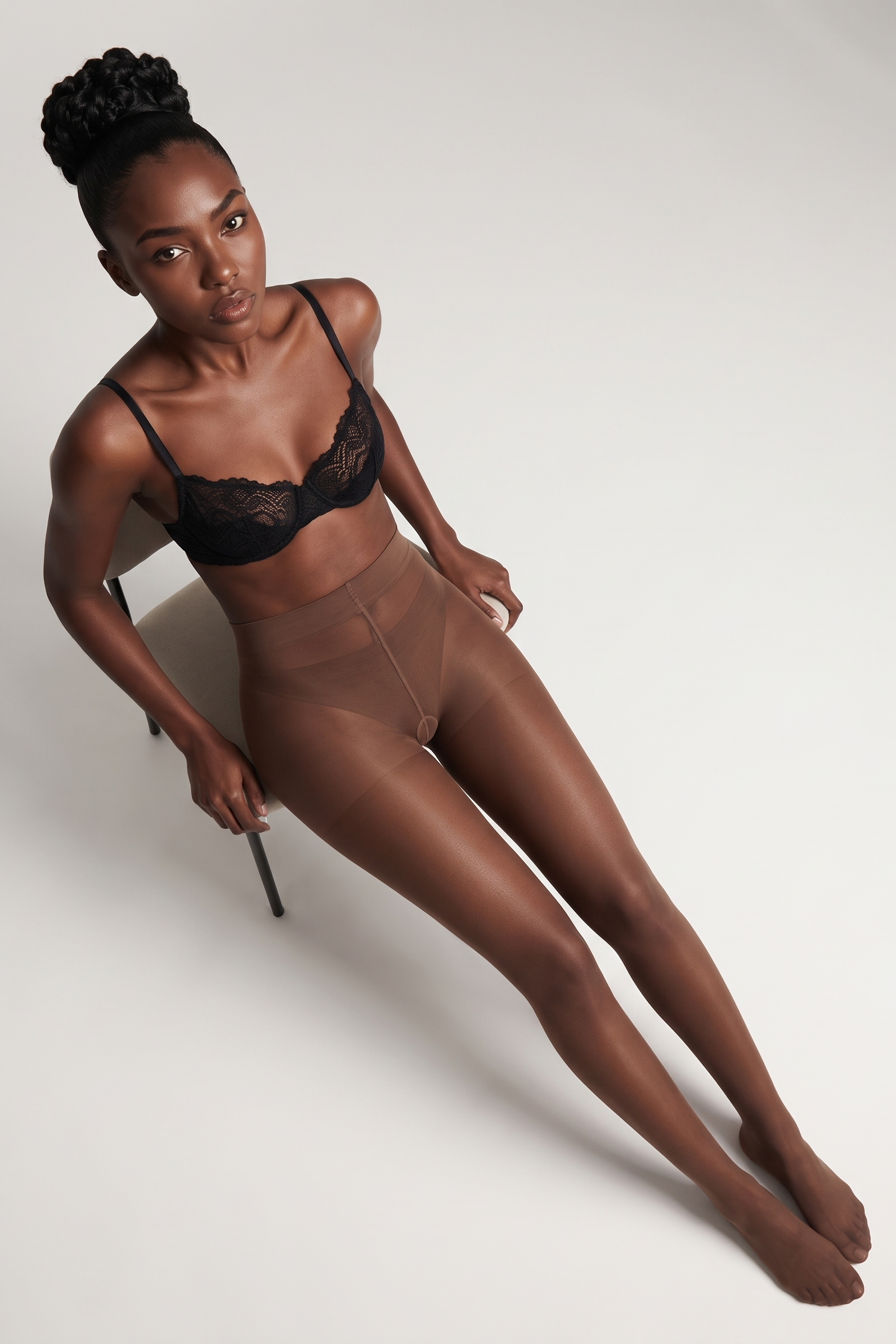 A model wearing sheer black lace underwire bras and matching sheer black pantyhose, posed on a minimalist white studio chair against a light gray background.