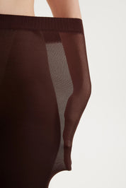 Close-up of a model wearing sheer dark brown tights or a pantyhose, showing the ribbed waistband and semi-transparent fabric panels against a plain light background.