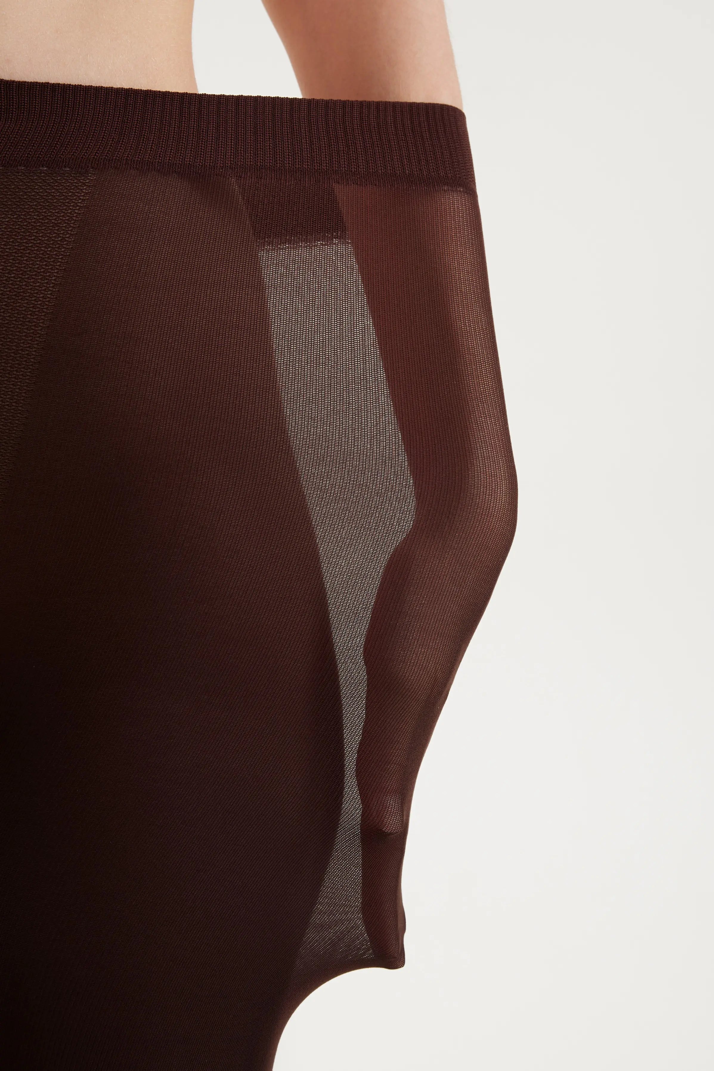 Close-up of a model wearing sheer dark brown tights or a pantyhose, showing the ribbed waistband and semi-transparent fabric panels against a plain light background.