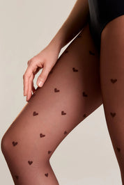 Close-up of a model's legs wearing sheer black tights with a scattered small heart pattern, paired with a black bodysuit against a plain light beige background.