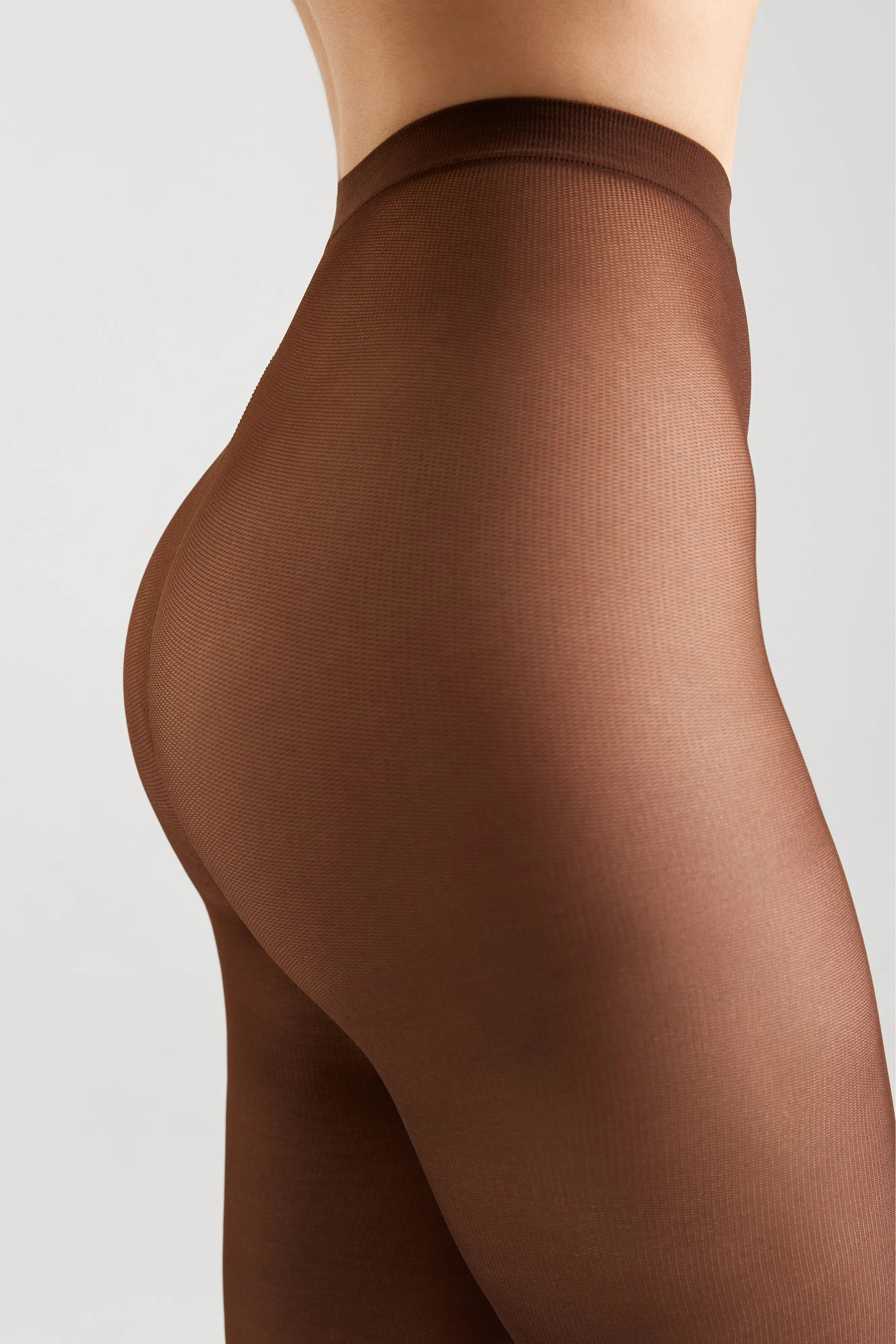 Close-up side view of a model wearing sheer brown hosiery against a light gray studio background.