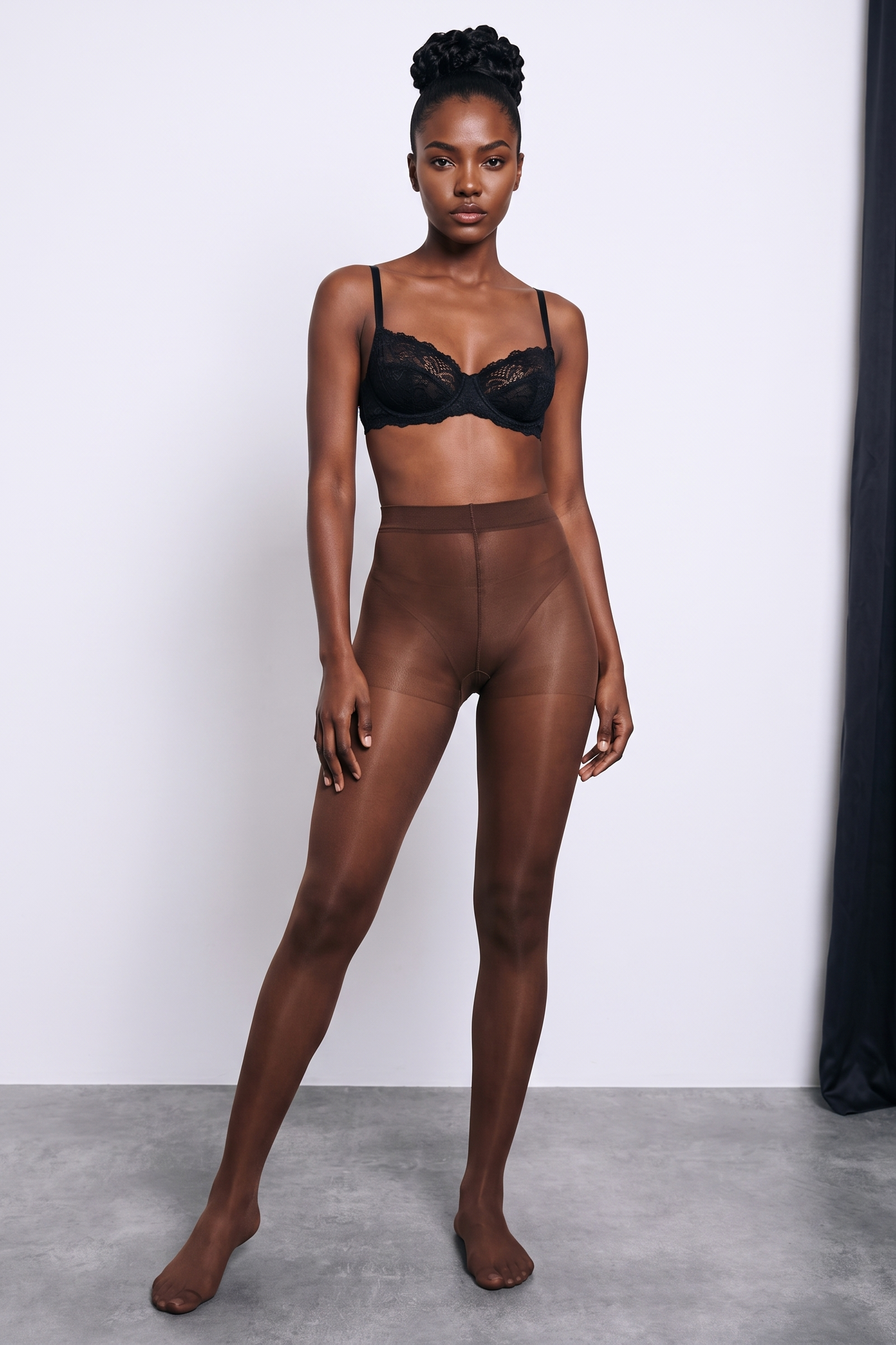 A model stands barefoot in a studio wearing sheer brown tights and a black lace bra against a light gray wall and concrete floor.